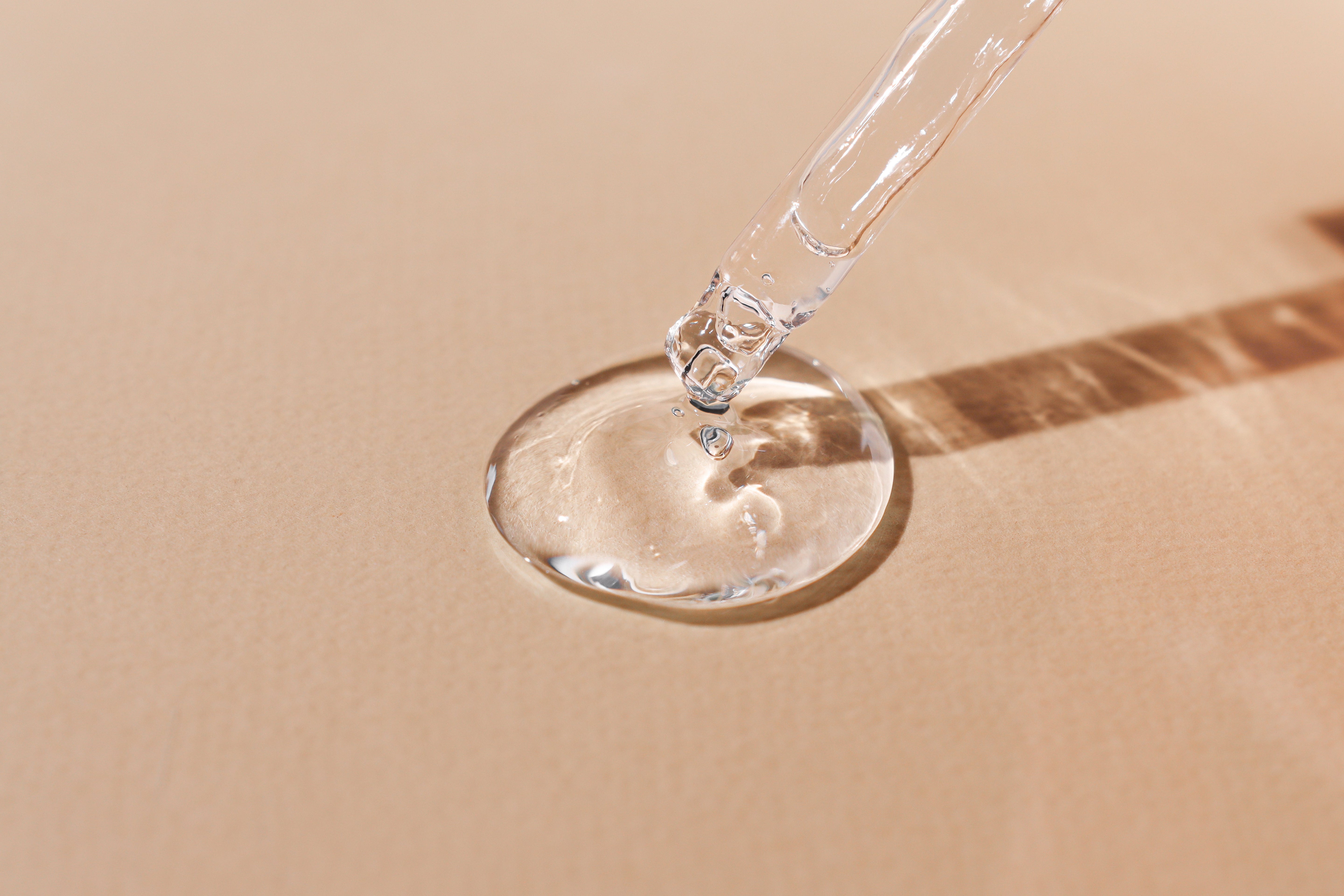 Clear serum dripping from a glass dropper onto a brown table – minimal skincare product photography.