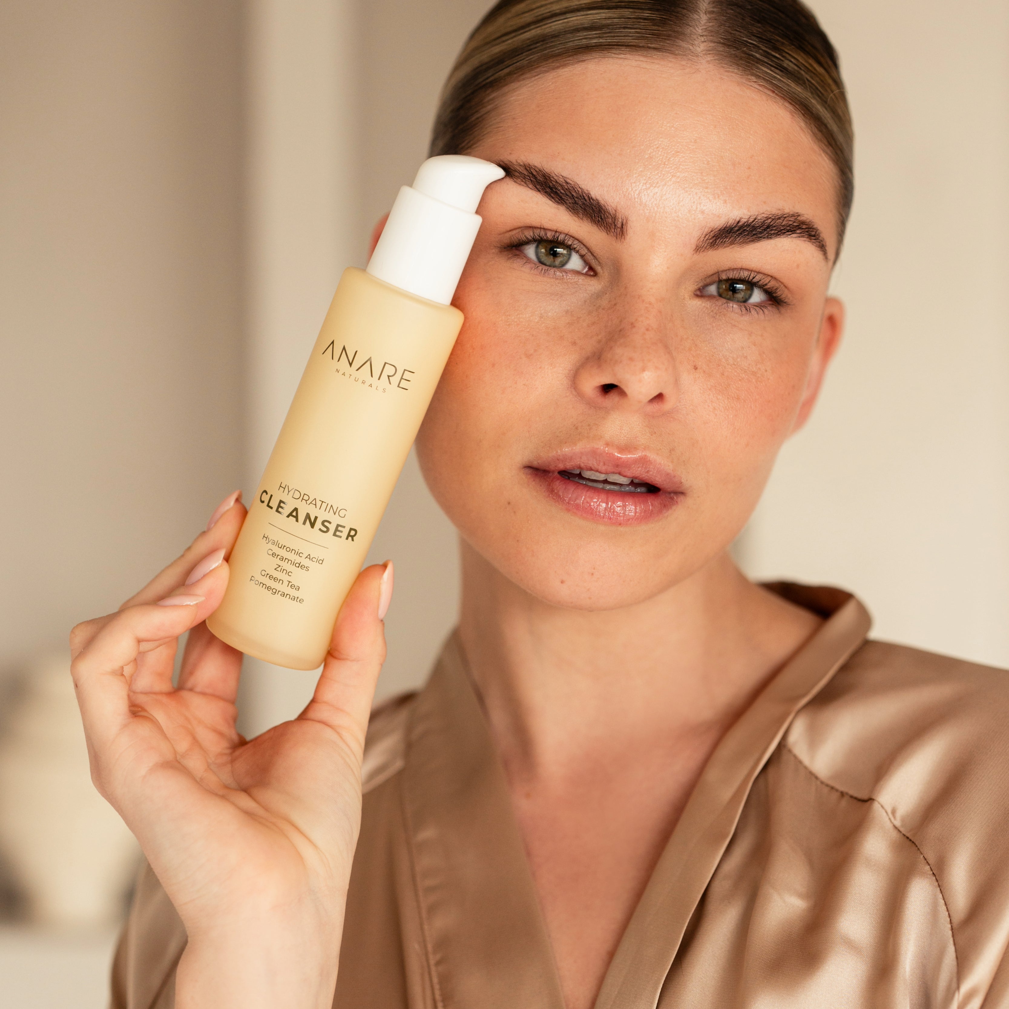A woman with dark blonde hair holding the Hydrating Cleanser next to her head, highlighting the product's sleek design and cleansing benefits.